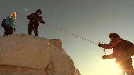 team of climbers climbs a mountain on a rope. free woman helps a male traveler climb a mountain. teamwork of tourists. Travel and adventure in mountains at sunset. businessmen insure each other.の写真素材