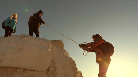 team of climbers climbs a mountain on a rope. free woman helps a male traveler climb a mountain. teamwork of tourists. Travel and adventure in mountains at sunset. businessmen insure each other.の写真素材