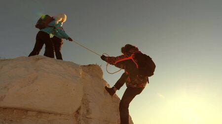 team of climbers climbs a mountain on a rope. free woman helps a male traveler climb a mountain. teamwork of tourists. Travel and adventure in mountains at sunset. businessmen insure each other.の写真素材
