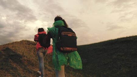 strong and healthy girls travelers travel in colorful raincoats, climb mountain, hold hands, help each other. Free women tourists with backpacks on rise to top of hill, rejoice in victory and jumpの写真素材