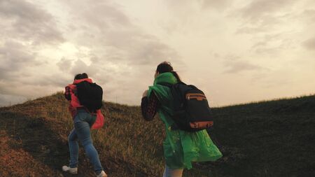 strong and healthy girls travelers travel in colorful raincoats, climb mountain, hold hands, help each other. Free women tourists with backpacks on rise to top of hill, rejoice in victory and jumpの写真素材