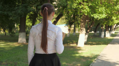 A student walks down the street with textbooks in her hands in the summer park. schoolgirl in the city. girl teenager hurries to school with books.の写真素材