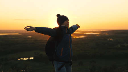 free girl tourist travels in nature alone. young woman traveler with a backpack comes to edge of the peak of sunrise, raises her hands in air and spins, enjoys victory, beautiful dawn and landscape.の写真素材
