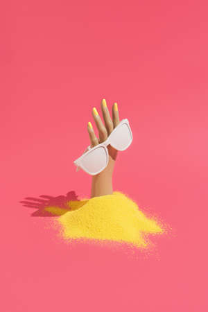 Plastic womans hand coming out of sand, holding sunglasses on intensive pink background. Minimal summer aesthetic.の写真素材