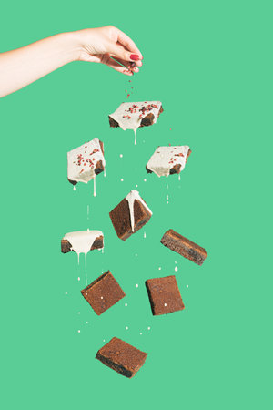 Minimal composition with chocolate brownies covered
with white cream floating against green background. Female hand pouring crumbs on cookies. Tasty dessert concept. Abstract food visual.の写真素材