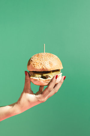 Modern scene with womans hand holding a burger against green background. Minimal composition with copy space. Fast food and street food concept.の写真素材