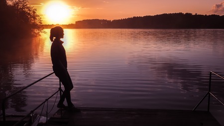 Silhouette of a girl on the background of the sun. Girl standing near water outdoors. Gold sunset lake. Young woman thinking about something river during golden sunset. Backlight photo shotの写真素材