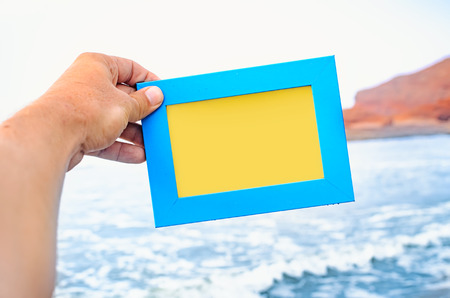 A male hand holds a blue photo frame with empty space seascape on background. Travel concept.の写真素材