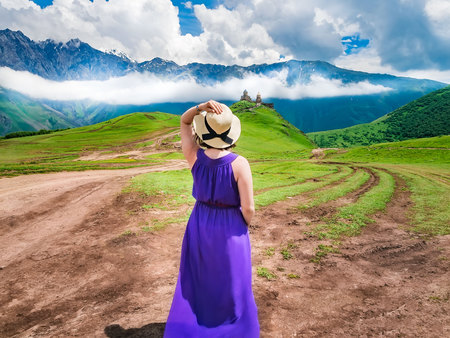 The scenic view from Kazbegi Georgia. Girl in long dress and hat stands and looks at the church of the Holy Trinity in Stepantsmindaの写真素材