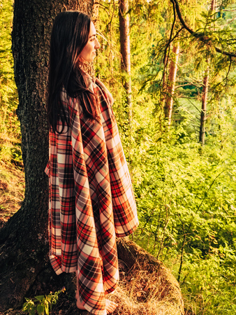 Beautiful caucasian brunette girl wrapped in a checkered plaid in the forest at sunset standing near a tree. The girl leans on a tree in the forest admiring the sunset on nature. Camping lifestyle. Unity with nature conceptの写真素材