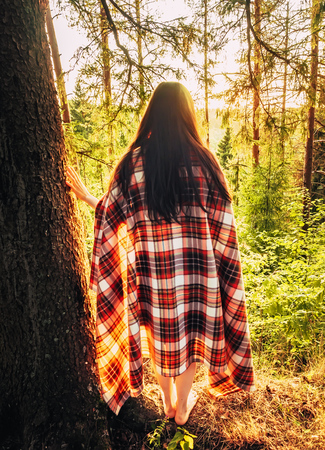 Beautiful caucasian brunette girl wrapped in a checkered plaid in the forest at sunset standing near a tree. The girl leans on a tree in the forest admiring the sunset on nature. Camping lifestyle. Unity with nature conceptの写真素材
