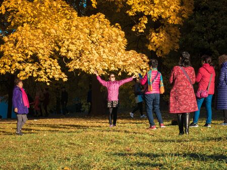 Moscow, Russia - October 11, 2018: Chinese tourists walks autumn park. Asian people take pictures on the background of a beautiful yellowed maple in the autumn park. Sunny day in the city park Izmailovo.のeditorial素材