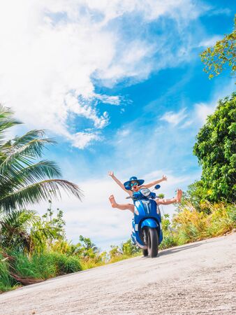Crazy funny woman with flying hair riding a motorbike on a blue sky and green tropics background. Young bizarre girl with dark hair in sunglasses on a blue scooter in vintage style racing downhill. Concept of summer holidays and vacation adventures.の写真素材