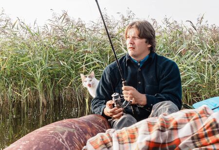 Fisherman catches fish from a boat on beautiful small river in the early morning at sunrise with his cat.の写真素材