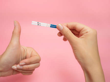 Female hand holding positive pregnancy test and Showing thumb up gesture isolated on pink...