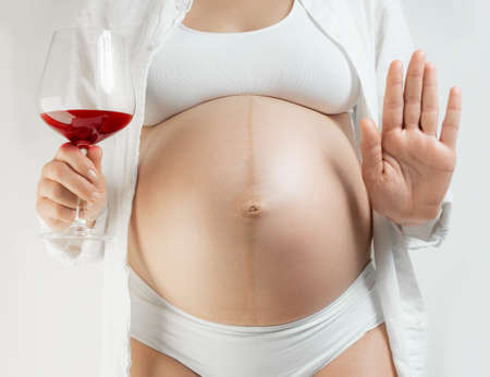 Lady in the third trimester of pregnancy informs that you should not drink alcohol during pregnancy. A pregnant young woman with a glass of red wine in hand and a Stop gesture against a white background. The girl in the last month of pregnancy in white underwear. Alcohol and childbearing concept.の写真素材