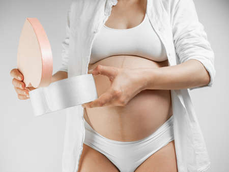 Lady in the third trimester of pregnancy. A pregnant young woman in white shirt opens a gift box white background. A girl in the last month of pregnancy opens a pink box and looks what's inside. Preparation for motherhood.の写真素材