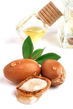 Three argan nuts with green leaves glass bottles on isolated white background. Chopped argan nut with a drop of oil. Whole and half Moroccan Argania Spinosa seeds for the production of oilの写真素材