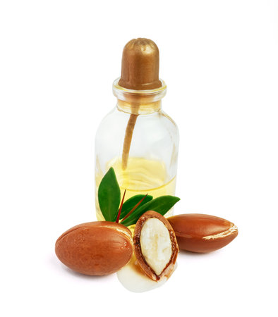 Three argan nuts with green leaves glass bottles on isolated white background. Chopped argan nut with a drop of oil. Whole and half Moroccan Argania Spinosa seeds for the production of oilの写真素材