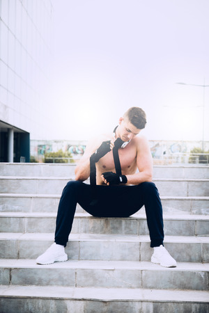 Young male preparing for hard workout and putting his gloves for training. Wearing tracksuit without shirt. In the background modern sport center. Selective focus.の写真素材