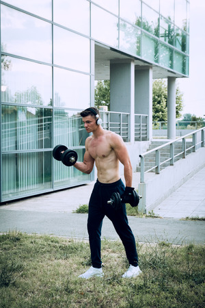 Young man listening to the music and working out with dumbbells in front of the modern sport center. Wearing tracksuits without shirt. Selective focusの写真素材