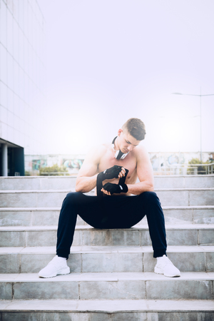Young male preparing for hard workout and putting his gloves for training. Wearing tracksuit without shirt. In the background modern sport center. Selective focus.の写真素材