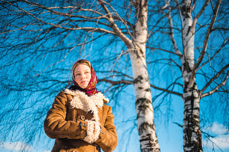 Beautiful blond woman in russian winter suit stay near the birch and thoughtfully looks afar. Girl clothed scarf, sheepskin coat.の写真素材