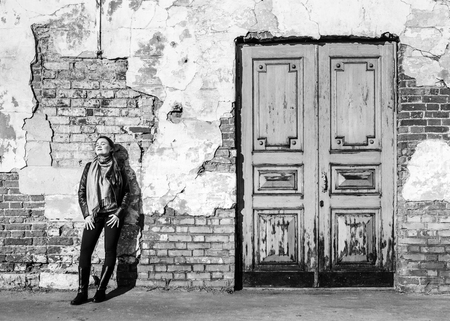 Young girl, wearing leather jacket and scarf, enjoys spring sun rays near texture ancient brick wall and doors.の写真素材