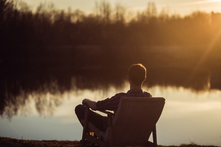 Man in the seat at bank of  lake also looks afar on sunset. Relaxing and freelance concept.の写真素材