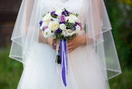 Young beautiful Bride holding bouquet of purple, violet, yellow, and cream-colored roses, and other flowers mixed with wild greensの写真素材
