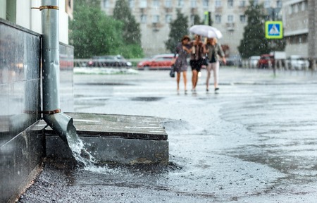 Rain water flowing from metal downspout during a flood. Three women cross the road at backgrond bokeh. Concept of protection against heavy downpour , rainy weatherの写真素材