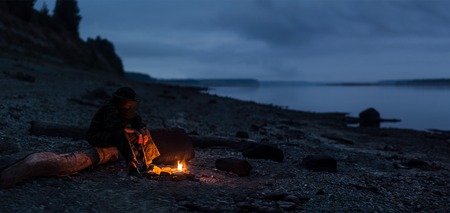 Hiker sitting by the bonfire on river shore. Focus on the bonfire and feetの写真素材