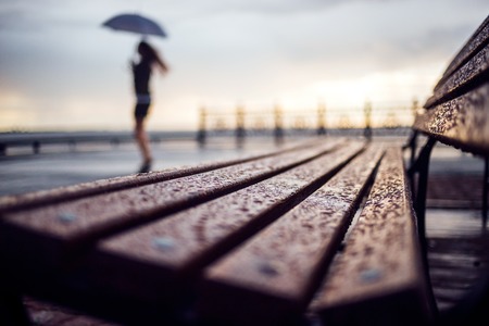 Wet bench and silhouette woman with umbrela. Rainy weather concept