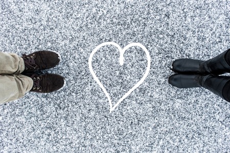 Male and Female boots standing at heart symbol on asphalt covered gritty snow surface. Rough snowy. Cold Winter. Top view. Relations conceptの写真素材