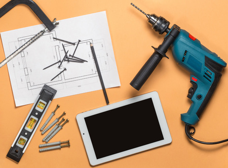 Set of construction tools to repair and white tablet on orange-brown table: drill, hammer, pliers,...