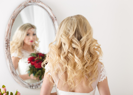 Beautiful curly haired bride in a wedding dress at a mirror in white room. Girl repeats the hairstyle and .の写真素材