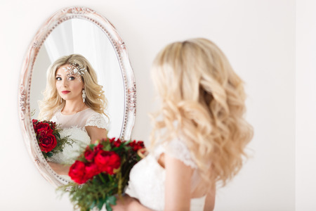Beautiful bride in a wedding dress at a mirror in white room. Girl repeats the hairstyle and makeup.の写真素材