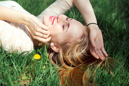 Blue-eyed Blonde Lying on the Grass with Dandelions. Cute Young Girfriend Lovely Laughing in Warm Sunny Day.の写真素材