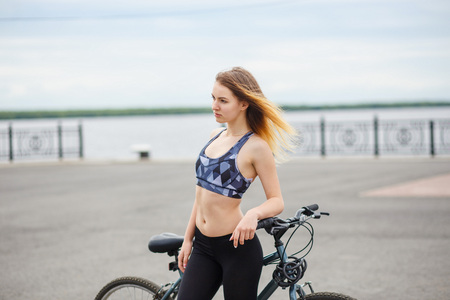 Smiling slim young woman standing with bicycle on embankment and looking at camera. Girl dressed in a sports top and leggings.の写真素材