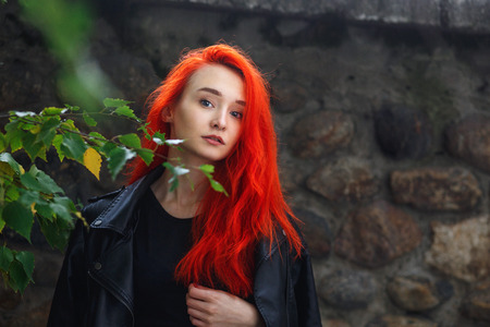 Portrait of beautiful redhead girl on dark wall outdoor. Unformal woman with vibrant colorful hairstyle standing among birch branches. Autumn time.の写真素材