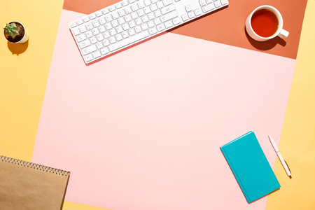 Trendy home office workspace. Flat lay composition of keyboard, cactus, diary with pen and cup of tea on colorful desk. Pink, yellow, aquamarine and and brown colours. Blogger's home office.の写真素材