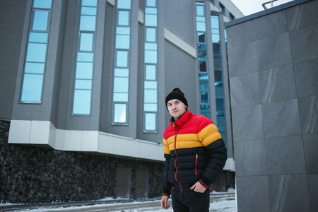 Handsome young man outdoor in colorful winter jacket looking away at cold gray urban backgroundの写真素材