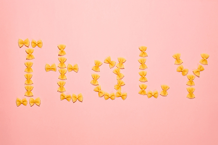Lettering Italy made up of farfalle pasta on pink background. Flat lay composition for bloggers, articles, magazines.の写真素材