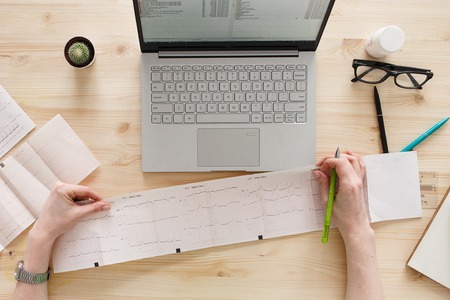 Professional doctor cardiologist analyzing a cardiogram of patients and writing medical records on computer and medical equipment all around, desktop top viewの写真素材