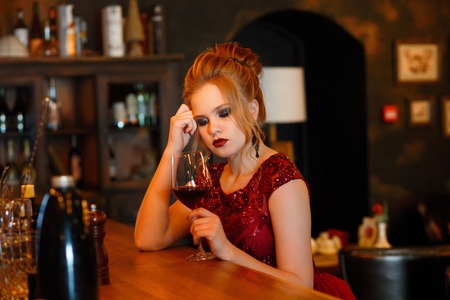Portrait of gorgeous beauty young blonde woman in red dress sitting at the bar with glass of red wine in luxury interiorの写真素材
