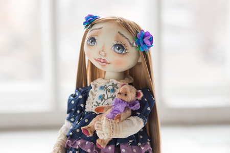 Portrait of textile handmade vintage doll with blue eyes, long brown hair in old blue textile dress with gentle print, in white shirt with embroidery white boots on white background. She holding little textile brown bearの写真素材