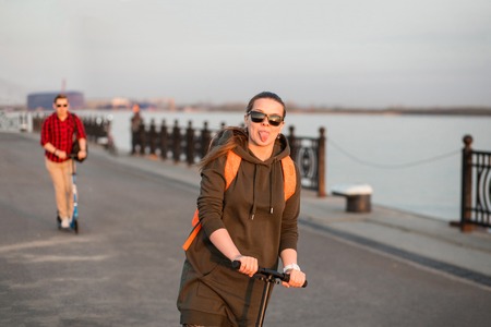 Cheerful friends riding kick scooter. Girl wearing dark green hoody with orange backpack and man wearing red shirt on background. Happy friends having fun in weekendの写真素材