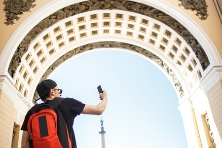 Young guy tourist with backpack taking mobile photo of historical archway in the palace square in St. Petersburgの写真素材