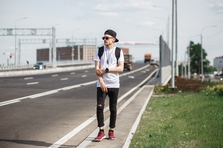 Stylish man hitchhiker on the street or motorway. He wearing in black jeans, black T-shirt, black cap with round sunglassesの写真素材