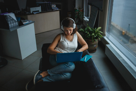 Young woman listening music in headphones and working on laptop on the sofa in office. Recreation,...